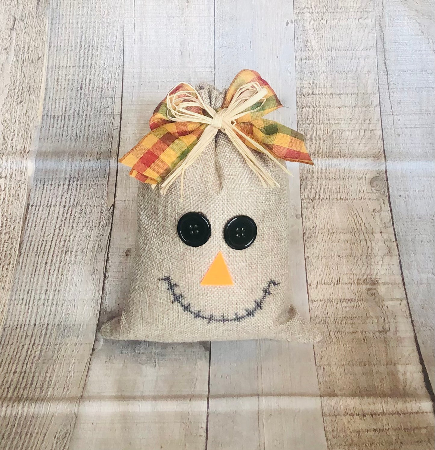Burlap Bag Scarecrow Craft Kit from Seniorly Creations. This is a picture of the completed craft with plaid and raffia bow at the top, buttons for eyes, felt for nose. 